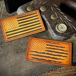 May include: Two wooden American flag patches with a distressed design. The patches feature the stars and stripes in a burnt orange and black color scheme. The patches are rectangular and appear to be laser cut. The background includes a vintage saw.