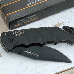 May include: Black pocket knife with a black handle and a silver blade. The knife has a safety feature and a belt clip. The knife is engraved with the text "TRACY #997" and "TAC-FORCE".