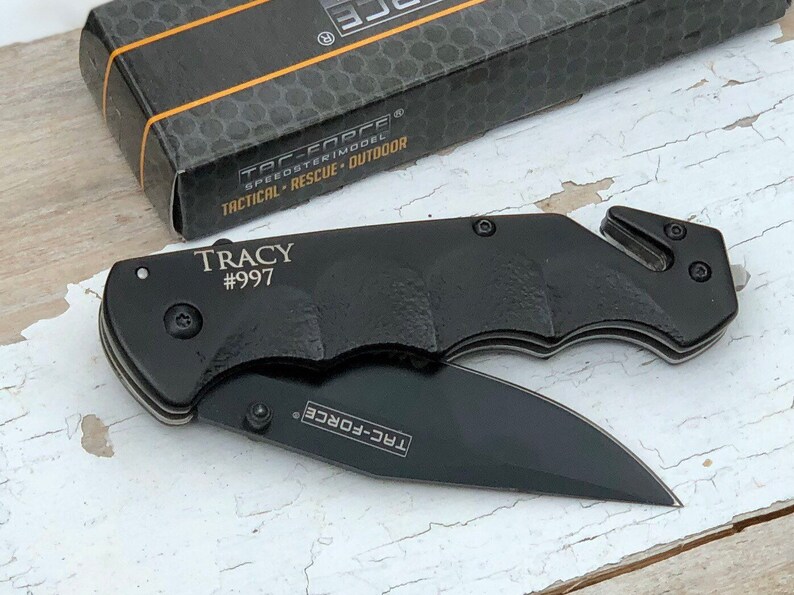 May include: Black pocket knife with a black handle and a silver blade. The knife has a safety feature and a belt clip. The knife is engraved with the text "TRACY #997" and "TAC-FORCE".