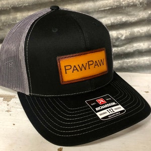 May include: Black and gray trucker hat with a brown leather patch that says "PAWPAW" on it. The hat is made by Richardson 112.