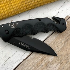 May include: Black folding knife with a serrated blade and a safety feature. The knife has a black handle with a textured grip and the words "TRC-FORCE" and "TRACY #997" engraved on it.