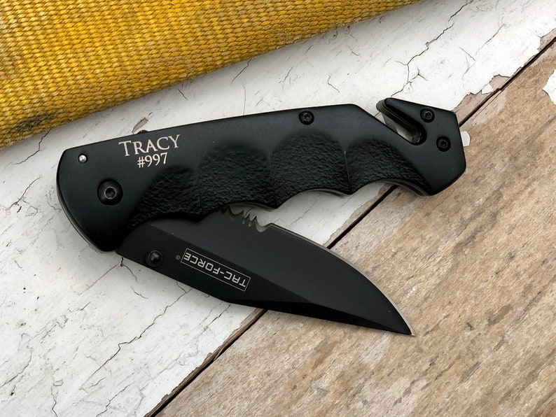 May include: Black folding knife with a serrated blade and a safety feature. The knife has a black handle with a textured grip and the words "TRC-FORCE" and "TRACY #997" engraved on it.