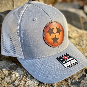 May include: A gray and white trucker hat with a brown leather patch featuring three black stars. The patch is a stylized version of the state of Tennessee's flag. The hat has an adjustable strap with a black label that reads "Richardson 112 Original Trucker Adjustable".