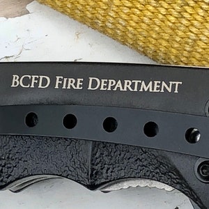 May include: Black folding knife with a black handle and a silver blade. The knife has a pocket clip and a row of holes on the handle. The knife is engraved with the text "BCFD FIRE DEPARTMENT".