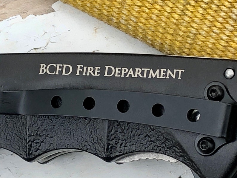 May include: Black folding knife with a black handle and a silver blade. The knife has a pocket clip and a row of holes on the handle. The knife is engraved with the text "BCFD FIRE DEPARTMENT".