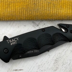 May include: Black folding knife with a safety blade and a silver inscription that reads "TRACY #997".