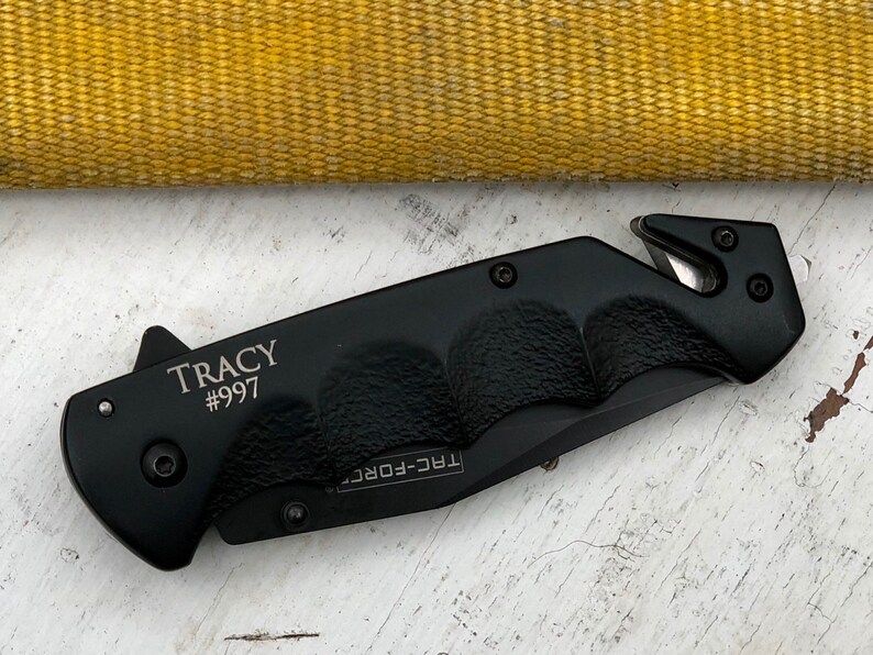 May include: Black folding knife with a safety blade and a silver inscription that reads "TRACY #997".