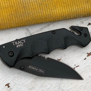 May include: Black folding knife with a serrated blade and a safety feature. The knife has a black handle with a textured grip and the words "TRAC-FORCE" and "TRACY #997" engraved on it.