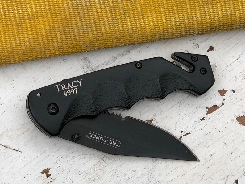 May include: Black folding knife with a serrated blade and a safety feature. The knife has a black handle with a textured grip and the words "TRAC-FORCE" and "TRACY #997" engraved on it.