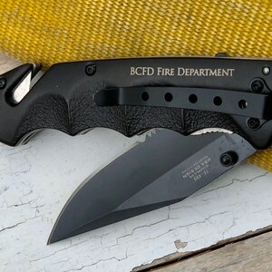 May include: Black folding knife with a black handle and a partially open blade. The handle has a pocket clip and the text "BCFD FIRE DEPARTMENT" is engraved on the side.