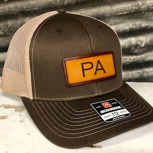 May include: A brown trucker hat with a tan mesh back and a curved brim. The hat features a rectangular leather patch with the letters "PA" in a bold font. The hat has a Richardson 112 Trucker tag.