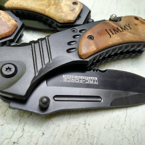 May include: A black folding knife with a wood handle. The handle is engraved with the name "JIMMY". The blade is partially open and has the text "TRC-FORCE SPEEDSYSTEM MODEL" engraved on it.