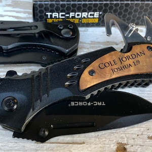 May include: A black pocket knife with a wood handle engraved with the text "COLE JORDAN JOSHUA 1:9". The knife has a black blade and a silver hook.