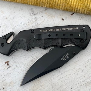 May include: Black folding knife with a serrated blade and a safety feature. The knife has a black handle with a textured grip and the words "Shelbyville Fire Department" engraved on the side.