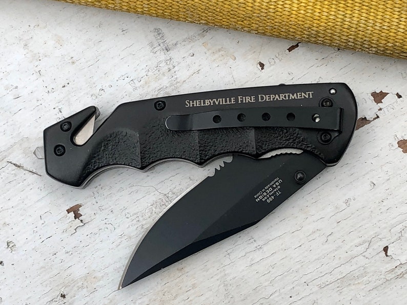 May include: Black folding knife with a serrated blade and a safety feature. The knife has a black handle with a textured grip and the words "Shelbyville Fire Department" engraved on the side.