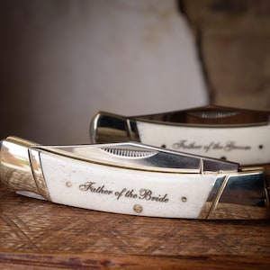 May include: Two folding pocket knives with metallic blades and off-white handles. The front knife is engraved with the text Father of the Bride, while the knife positioned behind it is engraved with the text Father of the Groom in script font.
