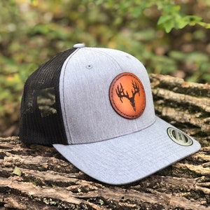 May include: A gray and black trucker hat with a brown leather patch featuring a deer skull with antlers. The patch is circular and has a stitched edge.