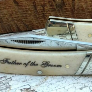 May include: Two pocket knives with bone handles and silver accents. The top knife is engraved with "Father of the Bride" and the bottom knife is engraved with "Father of the Groom".