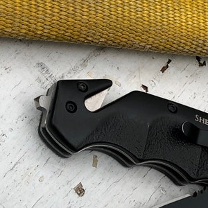 May include: Black folding knife with a serrated blade and a safety feature. The knife has a black handle and a silver blade.