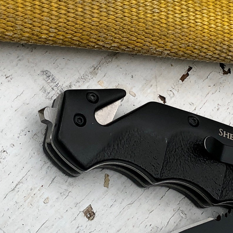 May include: Black folding knife with a serrated blade and a safety feature. The knife has a black handle and a silver blade.