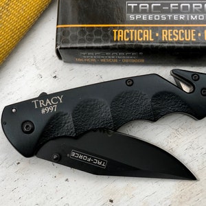 May include: Black pocket knife with a safety blade and a black handle. The knife has the text "TRACY #997" and "TAC-FORCE" engraved on it.
