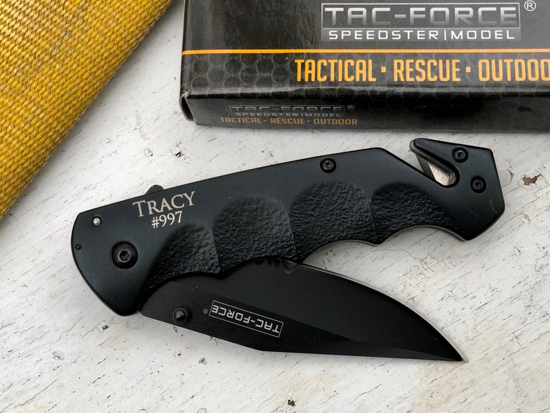 May include: Black pocket knife with a safety blade and a black handle. The knife has the text "TRACY #997" and "TAC-FORCE" engraved on it.