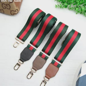 Op de afbeelding: Drie verstelbare tassenriemen met een groen en rood gestreept patroon. Elke riem is voorzien van goudkleurige metalen karabijnhaken en een lederachtig bevestigingspunt in zwart, donkerbruin of lichtbruin. Tekst: The bag is not for sale.