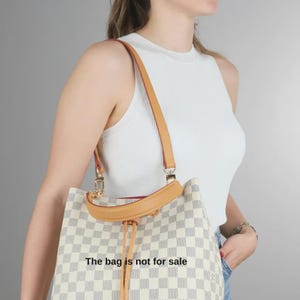 Puede incluir: Un bolso tipo cubo con un patrón de cuadros blancos y gris claro, ribete de cuero color canela, cierre de cordón y correa para el hombro. El bolso tiene herrajes dorados y el texto The bag is not for sale impreso al frente.