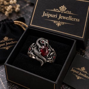 May include: A silver ring featuring a coiled snake design with a large oval red gemstone in the centre and small red stone accents. The ring is presented in a black box with the gold text Jaipuri Jewellerss printed on the lid and interior.