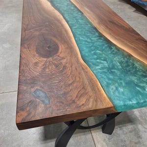 May include: A rectangular dining table featuring a live edge walnut wood slab with a prominent central knot and a translucent, swirling turquoise epoxy resin river inlay, supported by a black metal base with curved legs.