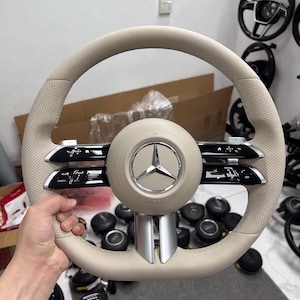 Könnte beinhalten: Ein beigefarbenes Lederlenkrad mit einer zentralen runden Nabe und einem verchromten Mercedes-Benz-Stern-Emblem. Das Lenkrad verfügt über perforierte Ledergriffe, schwarze Bedienfelder mit silbernen Akzenten und eine silberne vertikale Speiche.