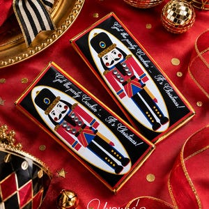 May include: Two rectangular chocolate bars wrapped in black paper featuring a colorful nutcracker illustration with gold accents and clear rhinestones. The text reads, Get the party Crackin'... It's Christmas! The bars have gold foil edges.