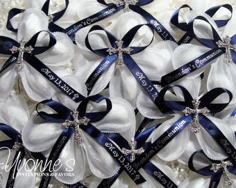May include: White flower-shaped favors with navy blue ribbons and silver cross charms. The ribbons are printed with "Brandon's Communion" and "May 13, 2017". The favors are for a religious event.