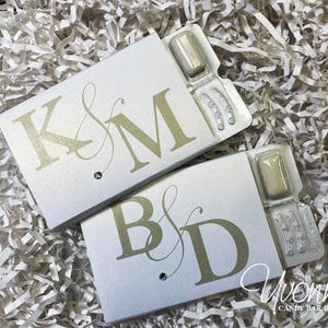 May include: Two personalized white favor boxes with gold initials and a small jewel, each containing gum. The boxes are on a bed of white shredded paper. The text "Yvonne's Candy Bar Boutique" is visible.