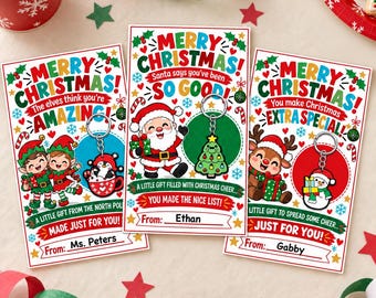 Könnte beinhalten: Drei rechteckige Weihnachtsgeschenkkarten mit farbenfrohen Feiertagsgrafiken und befestigten Metallschlüsselanhängern. Die Karten zeigen Illustrationen von Elfen, dem Weihnachtsmann und einem Rentier sowie Text wie Merry Christmas.
