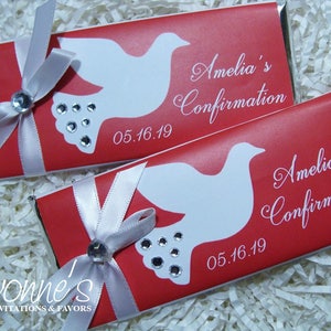 May include: Two red chocolate bar wrappers with white dove designs, text that reads "Amelia's Confirmation" and the date "05.16.19". Each wrapper is adorned with a silver ribbon and a rhinestone embellishment. The wrappers are from Yvonne's Invitations & Favors.