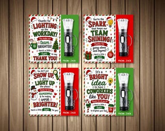 May include: Four rectangular cards with Christmas-themed graphics and text, each featuring a small black flashlight attached to the right side. The cards include phrases like Thanks for lighting up the workday and You're the spark that keeps our team shining, all signed From: Jack.