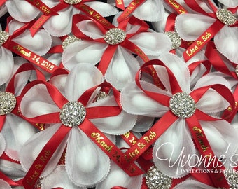 May include: Red and white favor boxes with a white satin bow and a rhinestone embellishment. The boxes are tied with a red satin ribbon with gold lettering that reads "March 24, 2018" and "Lisa & Rob".