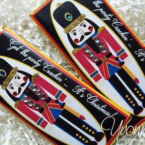 May include: Two chocolate bar wrappers with a Nutcracker design. The wrappers are black with gold glitter and feature a red and blue Nutcracker with a gold crown and a red nose. The text on the wrappers reads "Get the party crackin'... It's Christmas!" and "Yvonne's Invitations & Favors".