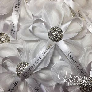May include: White satin ribbon favors with a silver rhinestone embellishment. The ribbon is personalized with the names "Charles and Denise" and the date "August 5, 2017".