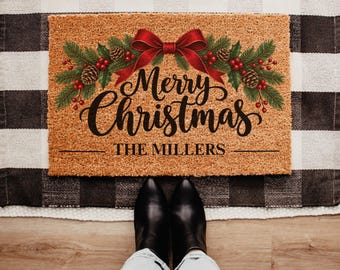 Op de afbeelding: Een rechthoekige beige kokosmat met een feestelijk ontwerp van groene dennentakken, rode bessen, dennenappels en een grote rode strik. De tekst Merry Christmas is in zwarte letters gedrukt, met daaronder de naam THE MILLERS in zwarte letters.