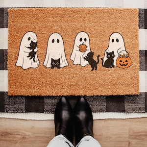 Peut inclure: Un paillasson rectangulaire en fibre de coco beige avec un motif imprimé représentant quatre fantômes blancs interagissant avec des chats noirs. Un fantôme porte un chat, un autre est près d'un chat, un troisième tient une pelote et le quatrième est assis à côté d'une citrouille.