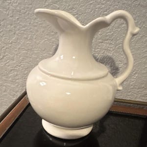May include: A glossy off-white ceramic pitcher featuring a rounded body, a wide base, and a curved handle. The vessel has a flared rim with a distinct pouring spout and a smooth, solid-colored finish throughout its entire exterior surface.