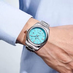 Könnte beinhalten: Eine Armbanduhr aus Edelstahl mit einem leuchtend türkisfarbenen Zifferblatt, silberfarbenen Stundenmarkierungen und leuchtenden Zeigern. Die Uhr verfügt über ein Datumsfenster auf der Drei-Uhr-Position und ein poliertes Metallgliederarmband.