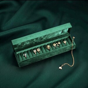 May include: A rectangular dark green velvet jewelry box with a hinged lid, containing five gold-toned rings with various stone settings and a delicate gold chain necklace draped over the right side of the velvet-lined interior storage slots.