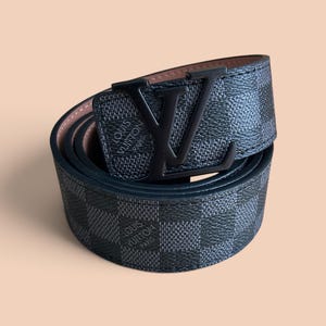 Puede incluir: Un cinturón enrollado con un patrón de cuadros gris oscuro y negro con el texto Louis Vuitton Paris impreso en la superficie. El cinturón incluye una hebilla de metal negro mate con forma de letras LV, con un forro interior de color canela.