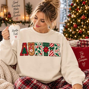 May include: A cream-coloured long-sleeve sweatshirt featuring the word AUNTIE in large, festive, patterned letters with Christmas tree, gingerbread, holly, poinsettia, and candy cane designs. A white mug with the text Auntie Fuel and a heart is held.