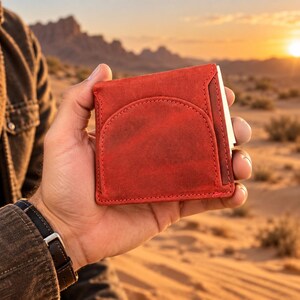 May include: A square red leather wallet featuring a semi-circular stitched pocket on the front face. The wallet has a matte, textured finish with visible stitching along the edges and a slim profile, held in a palm against a desert landscape.
