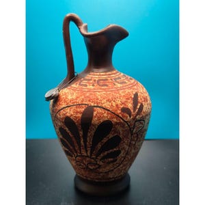 May include: A terracotta-colored ceramic pitcher featuring a dark brown handle and a flared rim. The body displays a mottled orange and brown texture with black painted palmette motifs and a geometric border pattern near the neck area.