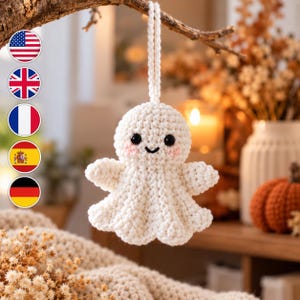 May include: A small, off-white crocheted ghost ornament featuring a rounded head, two black bead eyes, a stitched black smile, and soft pink blush on the cheeks. The ghost has a flared, skirt-like base and a braided yarn loop for hanging.